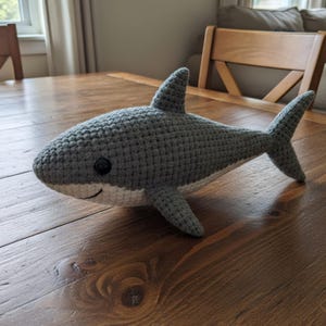 Peut inclure: Un jouet en forme de requin crocheté, gris et blanc, avec un visage souriant et des yeux noirs. Le requin fait à la main a des nageoires et une queue, et est posé sur une surface en bois. Mesure environ 30 cm.