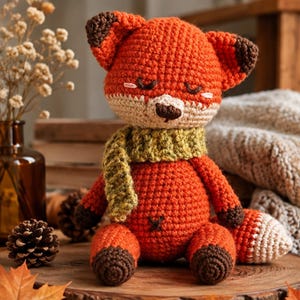 Puede incluir: Un peluche de zorro tejido a ganchillo a mano con hilo naranja brillante, patas y puntas de orejas marrón oscuro y un hocico color crema. El zorro lleva una bufanda texturizada de color verde oliva y tiene los ojos cerrados con detalles bordados.