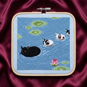 Peut inclure: Broderie au point de croix représentant une scène de bassin avec des chats. Les chats, noirs, blancs et calico, flottent sur l'eau avec des nénuphars et une fleur de lotus rose. L'œuvre est encadrée dans un cerceau en bois carré.