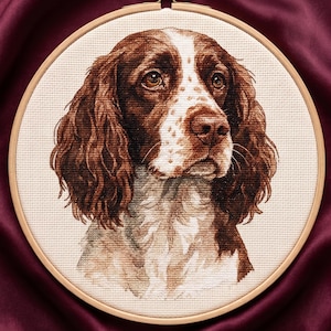 Peut inclure: Broderie au point de croix détaillée d'une tête de chien, avec des poils bruns et blancs, placée dans un cercle à broder en bois. Les yeux du chien sont bruns, et le fond est un tissu bordeaux foncé.