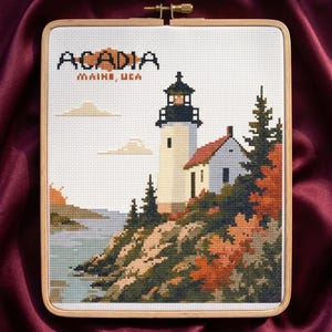 May include: A rectangular cross-stitch embroidery in a wooden hoop featuring a lighthouse on a rocky cliffside with trees and water. The text ACADIA MAINE, USA is stitched in black and orange. The scene includes a white lighthouse, a small house, and autumn-colored foliage in shades of orange, green, and brown.