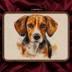 May include: A rectangular wooden embroidery hoop containing a cross-stitch portrait of a beagle dog. The dog features brown, black, and white fur with large floppy ears and dark eyes, rendered on a light cream-coloured fabric background.