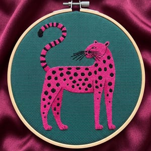 Puede incluir: Un leopardo rosa con manchas negras bordado sobre tela verde azulado, en un bastidor de madera. El leopardo tiene una cola larga, sobre un fondo de satén.