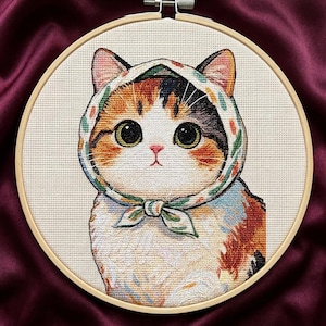 May include: A cross-stitch embroidery piece in a circular wooden hoop featuring a calico cat with large dark eyes wearing a white headscarf with a green and orange floral pattern tied under its chin against a cream-colored fabric background.