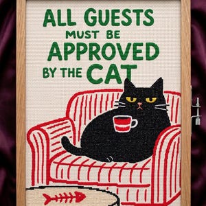 May include: Cross-stitch artwork in a wooden frame. The design features a black cat lounging on a red and white striped armchair, holding a mug. Text reads "ALL GUESTS MUST BE APPROVED BY THE CAT". A small table with a fishbone is in front.