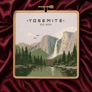 May include: A square wooden embroidery hoop containing a cross-stitch landscape of Yosemite, CA, USA. The design features a waterfall, mountains, a forest of green pine trees, and a reflective lake. Two small birds are stitched in the sky above.