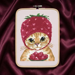 May include: A cross-stitch embroidery in a rectangular wooden hoop featuring a ginger tabby cat wearing a red strawberry-shaped hat with small pink hearts. The cat is holding a small red strawberry in its paws against a light pink fabric background.