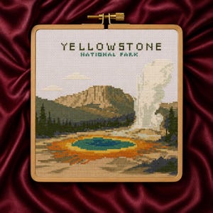 May include: A square wooden embroidery hoop containing a cross-stitch design of Yellowstone National Park. The scene features a geyser erupting, a vibrant blue and orange thermal pool, green pine trees, and mountains under the text Yellowstone National Park.