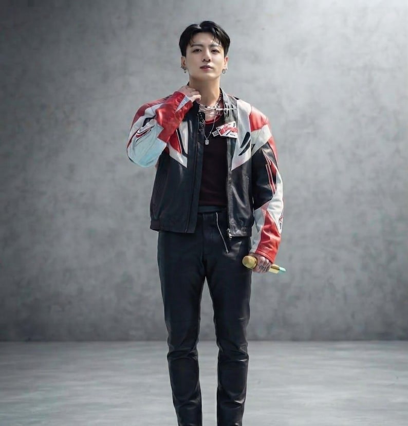 May include: A person wearing a black, red, and white leather racing jacket over a dark maroon shirt, paired with black leather trousers. The outfit includes a layered silver chain necklace and a gold-colored microphone held in one hand.