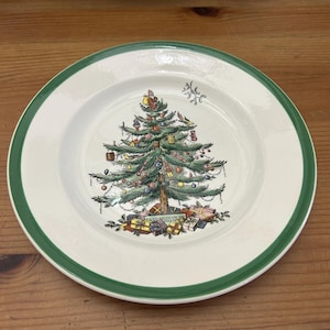 Puede incluir: Un plato de cerámica redondo con base color crema y borde verde oscuro. El centro presenta una ilustración detallada de un árbol de Navidad decorado con adornos coloridos, una estrella en la punta y regalos envueltos en la base.