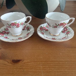 May include: Two white teacups and saucers with a floral rose pattern in pink and red. The rims and handles are trimmed with gold. The saucers have a scalloped edge.