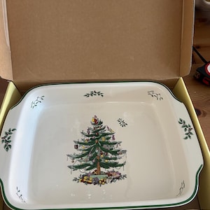 May include: A square white ceramic baking dish with rounded corners and a green rim. The centre features a detailed illustration of a decorated Christmas tree with gifts underneath. Small sprigs of holly are painted along the inner edges.