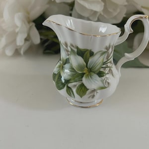 Peut inclure: Un pot à crème en porcelaine blanche avec un motif floral de fleurs blanches et de feuilles vertes. Le pot à crème a un bord et une anse dorés. Le fond présente des fleurs blanches.
