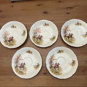 Può includere: Cinque piattini vintage color crema con un design a scena di caccia. Ogni piattino presenta un pozzetto centrale e un bordo decorativo. L'illustrazione raffigura un cavaliere a cavallo, un pescatore e un albero con fogliame autunnale.