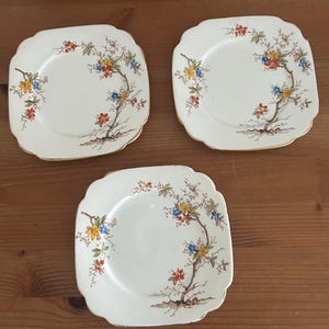 May include: Three square, off-white plates with scalloped edges. Each plate features a floral design with yellow, blue, and red flowers on a brown branch. The plates have a gold trim and are arranged on a wooden surface.