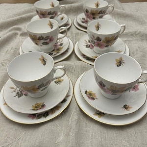 Peut inclure: Service à thé de six pièces, comprenant tasses, soucoupes et petites assiettes blanches. Chaque pièce est ornée d'un motif floral rose, jaune et vert, avec des bordures dorées. L'ensemble est disposé sur une nappe neutre.