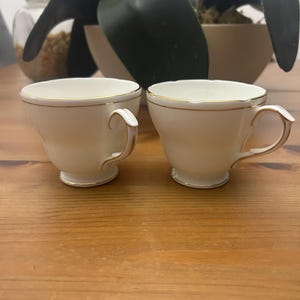 Könnte beinhalten: Zwei cremefarbene Teetassen mit goldenen Verzierungen und Henkeln stehen auf einer Holzoberfläche. Die Teetassen haben ein klassisches Design mit einem leicht gewellten Rand und einem kleinen Fuß. Eine dekorative Pflanze befindet sich im Hintergrund.