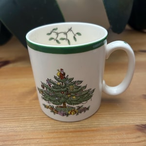 May include: A white ceramic mug featuring a green rim and a festive illustration of a decorated Christmas tree with a figure of Santa Claus at the top. The interior rim of the mug is decorated with a green mistletoe pattern and colourful gifts.