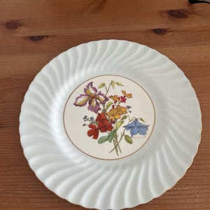 May include: White porcelain plate with a swirling rim design and a gold trim. The center features a floral bouquet with purple, red, yellow, and blue flowers. A decorative piece for serving.