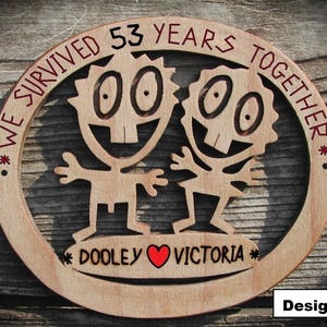 May include: A circular wooden plaque featuring a laser-cut design of two cartoon figures with large eyes and teeth. The text "WE SURVIVED 53 YEARS TOGETHER" is engraved along the top, and "DOOLEY" and "VICTORIA" with a red heart are at the bottom.