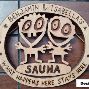 Op de afbeelding: Een rond houten bord met lasergesneden figuren van twee stripfiguren. De tekst luidt BENJAMIN & ISABELLA'S SAUNA WHAT HAPPENS HERE STAYS HERE. Het hout heeft een natuurlijke lichtbruine afwerking met donkere, ingebrande letters en details.