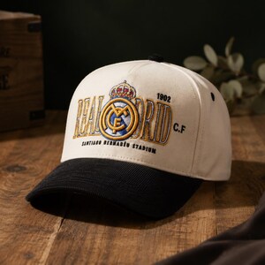 Pu&ograve; includere: Cappellino da baseball color crema con visiera nera. Presenta un logo ricamato del Real Madrid con corona, le parole "REAL MADRID" e "SANTIAGO BERNAB&Eacute;U STADIUM" in oro. L'anno "1902" &egrave; anch'esso ricamato.