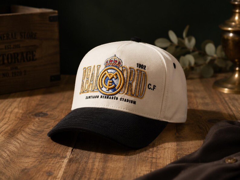 Pu&ograve; includere: Cappellino da baseball color crema con visiera nera. Presenta un logo ricamato del Real Madrid con corona, le parole "REAL MADRID" e "SANTIAGO BERNAB&Eacute;U STADIUM" in oro. L'anno "1902" &egrave; anch'esso ricamato.