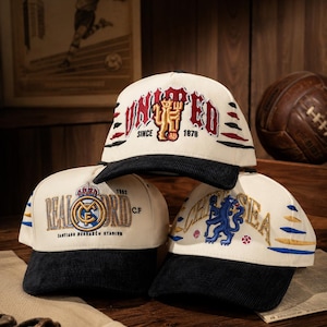 Pu&ograve; includere: Tre cappellini da baseball con loghi di squadre. I cappellini sono color crema con visiere nere e loghi ricamati. Un cappellino dice "UNITED" con "1878" e un logo. Un altro dice "REAL MADRID" e "1902". Il terzo dice "CHELSEA".