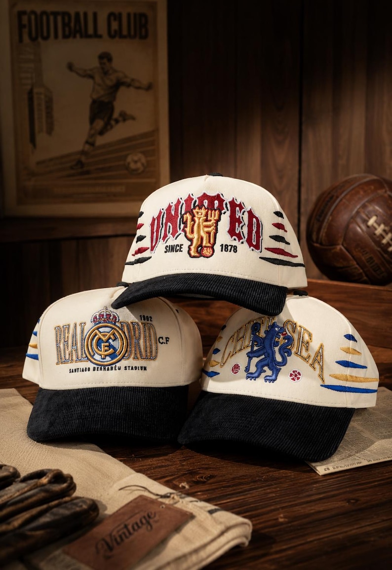 Pu&ograve; includere: Tre cappellini da baseball con loghi di squadre. I cappellini sono color crema con visiere nere e loghi ricamati. Un cappellino dice "UNITED" con "1878" e un logo. Un altro dice "REAL MADRID" e "1902". Il terzo dice "CHELSEA".