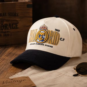 Pu&ograve; includere: Un cappellino da baseball bianco con visiera nera, con il logo del Real Madrid ricamato in oro, blu e bianco. Il cappellino include il testo "1902 C.F." e "Santiago Bernab&eacute;u Stadium".