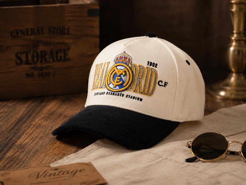 Pu&ograve; includere: Un cappellino da baseball bianco con visiera nera, con il logo del Real Madrid ricamato in oro, blu e bianco. Il cappellino include il testo "1902 C.F." e "Santiago Bernab&eacute;u Stadium".