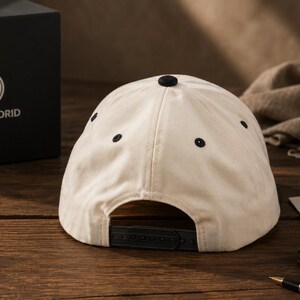Pu&ograve; includere: Cappellino da baseball color crema con dettagli neri, tra cui un bottone e occhielli. Chiusura snapback. Una scatola nera con il logo e il testo del Real Madrid &egrave; visibile sullo sfondo.