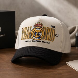 Pu&ograve; includere: Cappellino da baseball color crema e nero con il logo e il testo del Real Madrid ricamati in oro e blu. Il cappellino mostra "REAL MADRID" e "SANTIAGO BERNAB&Eacute;U STADIUM". Una scatola nera con il logo del Real Madrid &egrave; visibile sullo sfondo.