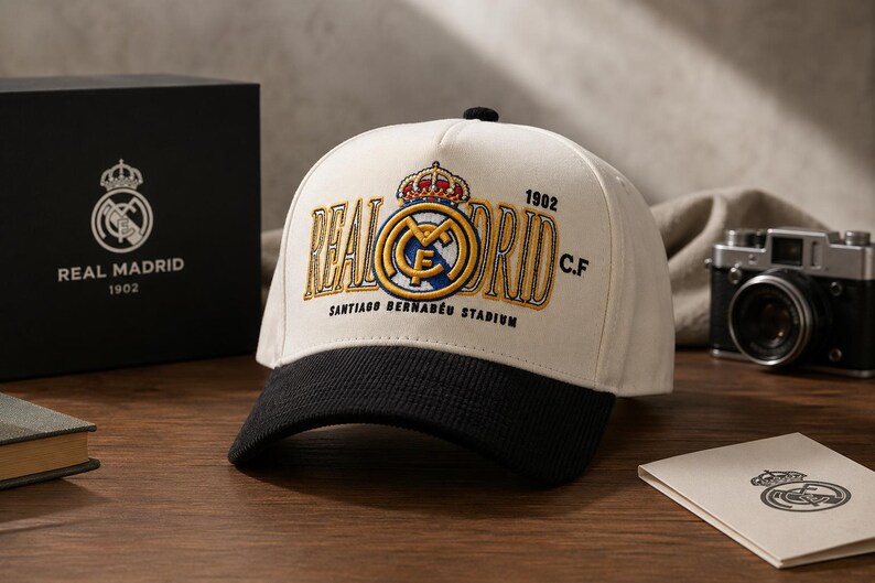 Pu&ograve; includere: Cappellino da baseball color crema e nero con il logo e il testo del Real Madrid ricamati in oro e blu. Il cappellino mostra "REAL MADRID" e "SANTIAGO BERNAB&Eacute;U STADIUM". Una scatola nera con il logo del Real Madrid &egrave; visibile sullo sfondo.