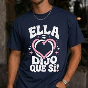 May include: Navy blue t-shirt with the text "ELLA DIJO QUE SÍ!" in white, featuring a pink and white heart design with a diamond. The shirt is a casual wear item.
