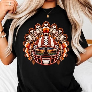 Op de afbeelding: Een zwart t-shirt met een afbeelding van een kalkoen die een footballhelm en een zonnebril draagt. De staartveren van de kalkoen hebben patronen van luipaardprint, ruitjes en strepen in oranje, bruin en wit.