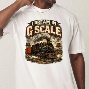 Può includere: T-shirt bianca con un'illustrazione vintage di un treno a vapore e la scritta "I Dream in G Scale". Il treno è nero e rosso, con fumo che esce dal camino. Il design ha un aspetto retrò e invecchiato.