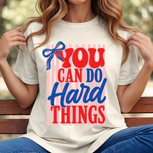 May include: A cream-colored short-sleeve t-shirt featuring a graphic print with vertical pink stripes. The text reads YOU CAN DO Hard THINGS in red and blue, accompanied by a blue ribbon illustration positioned above the word YOU.