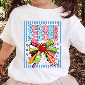 Può includere: Una t-shirt bianca con una stampa grafica che riporta il testo SECOND SECOND in blu e rosa. Il design include un grande fiocco colorato decorato con articoli scolastici come una mela rossa, matite gialle, un righello e un quaderno verde.