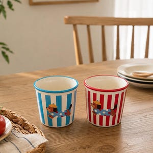 Puede incluir: Dos tazas de cerámica con diseño de rayas. Una taza tiene rayas azules y la otra rojas. Cada taza presenta un perro salchicha de dibujos animados con sombrero y atuendo de lunares.