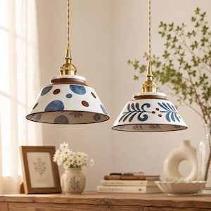 Op de afbeelding: Twee hanglampen met conische keramische kappen, opgehangen aan gedraaide stoffen snoeren met messing armaturen. De linker kap heeft blauwe en bruine stippen, terwijl de rechter kap een blauw bladpatroon op een witte achtergrond toont.