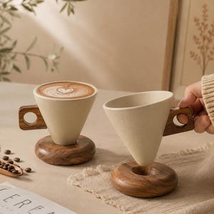Puede incluir: Dos tazas de café de cerámica color crema con asas y bases de madera. Una taza contiene café con arte latte. Las tazas están sobre una superficie beige con granos de café y un libro visible.