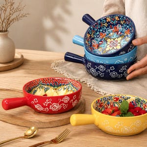May include: A selection of ceramic bowls with handles, featuring intricate floral designs in various colours. The bowls are filled with fruit and vegetables. A gold spoon and fork are on the wooden table. The bowls are in shades of blue, red, and yellow.