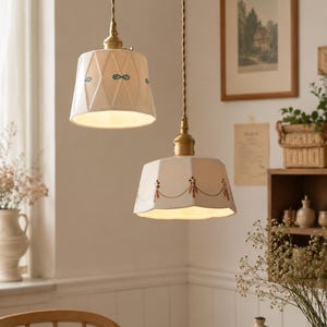 May include: Two pendant lights with cream-colored shades. The shades have decorative patterns and are suspended by twisted brown cords. The lights hang above a wooden table, creating a warm, inviting atmosphere. The fixtures have brass-colored hardware.