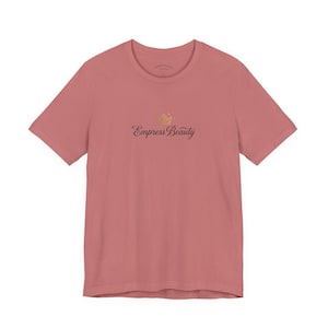 May include: A dusty rose-colored t-shirt with a crew neck. The shirt features the text "Empress Beauty" in a script font, with a gold crown graphic above the text. The shirt is made of a soft material.