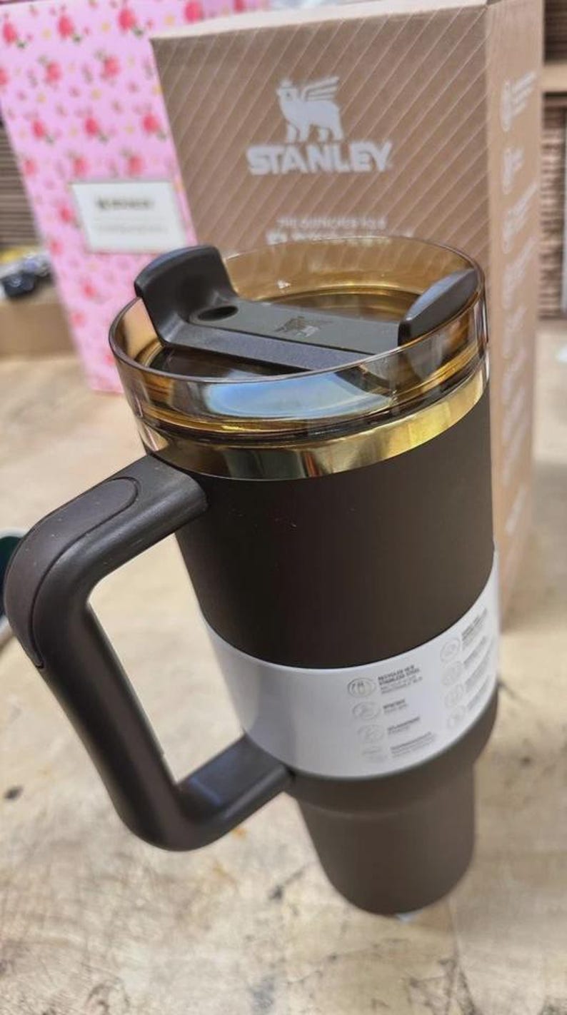 May include: A black and white Stanley travel mug with a handle. The mug has a gold-coloured rim and a clear lid. The Stanley logo is visible on the box behind the mug.