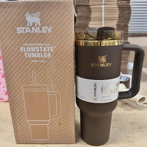 May include: A brown Stanley Flowstate tumbler with a handle and a gold-coloured rim, next to its cardboard box. The box features the Stanley logo and a drawing of the tumbler. The tumbler holds 1.18L.