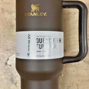 May include: A brown Stanley Quencher H2.0 Tumbler with a gold-coloured lid and handle. The tumbler has a white label with the text "QUENCHER H2.0 TUMBLER" and the Stanley logo. It holds 1.18 litres.