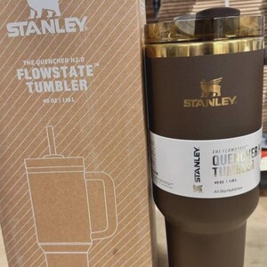 May include: A brown Stanley Flowstate tumbler with a gold rim and a straw, next to its cardboard box. The box features the Stanley logo and the text "The Quencher H2.0 Flowstate Tumbler 1.18 L". The tumbler is designed for all-day hydration.