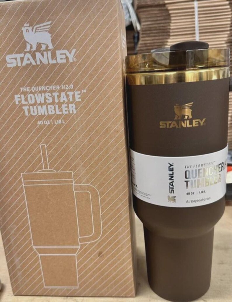 May include: A brown Stanley Flowstate tumbler with a gold rim and a straw, next to its cardboard box. The box features the Stanley logo and the text "The Quencher H2.0 Flowstate Tumbler 1.18 L". The tumbler is designed for all-day hydration.